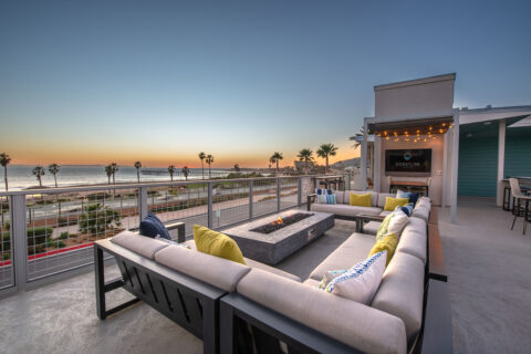 Outdoor sitting area with plush couch seating and fire pit, bistro seating and a flat screen TV, all overlooking evening ocean views